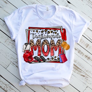 May include: White t-shirt with a colorful graphic that says "Taekwondo MOM." The design includes a leopard print background, red and gold medals, a karate uniform, and protective gear. The shirt is displayed on a white wooden surface.