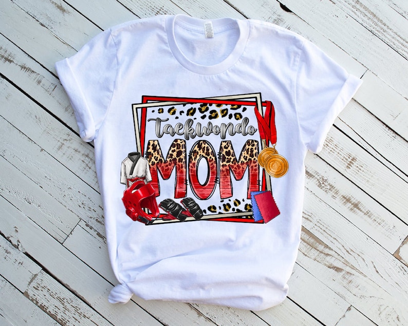 May include: White t-shirt with a colorful graphic that says "Taekwondo MOM." The design includes a leopard print background, red and gold medals, a karate uniform, and protective gear. The shirt is displayed on a white wooden surface.