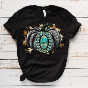 Puede incluir: Camiseta negra con un gr&aacute;fico de una calabaza gris con detalles turquesa y dorados sobre un fondo de estampado de leopardo marr&oacute;n y blanco.