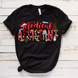 Puede incluir: Camiseta negra con un dise&ntilde;o navide&ntilde;o rojo y blanco que dice "Asistente M&eacute;dico". El dise&ntilde;o incluye bastones de caramelo, vendas y otros s&iacute;mbolos m&eacute;dicos.