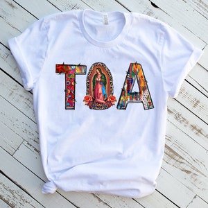 Puede incluir: Camiseta blanca con la palabra "TOA" impresa en letras de colores. Las letras est&aacute;n decoradas con un patr&oacute;n floral y una imagen religiosa de la Virgen Mar&iacute;a.