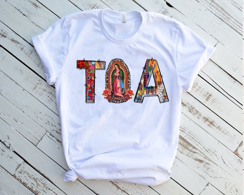 Puede incluir: Camiseta blanca con la palabra "TOA" impresa en letras de colores. Las letras est&aacute;n decoradas con un patr&oacute;n floral y una imagen religiosa de la Virgen Mar&iacute;a.