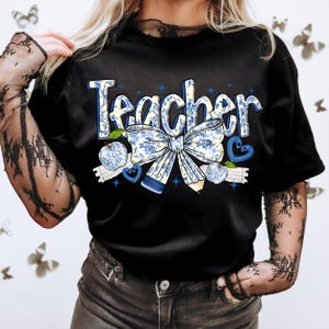 May include: Black t-shirt with the word "Teacher" in white and blue lettering with a blue and white floral bow and apples.