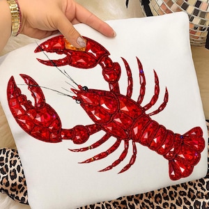 May include: A white cushion with a large, detailed red lobster design. The lobster is adorned with red gems, giving a sparkling appearance. The cushion is held by a hand, with a leopard print fabric visible in the background.