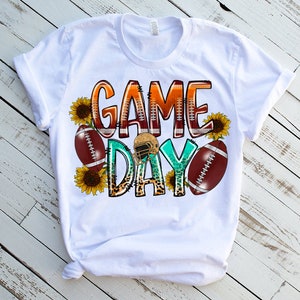 May include: White t-shirt with a graphic design that says "Game Day" in colorful letters with footballs and sunflowers.