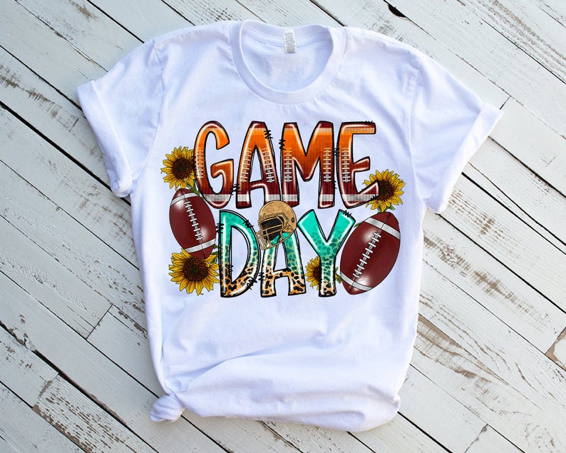 May include: White t-shirt with a graphic design that says "Game Day" in colorful letters with footballs and sunflowers.