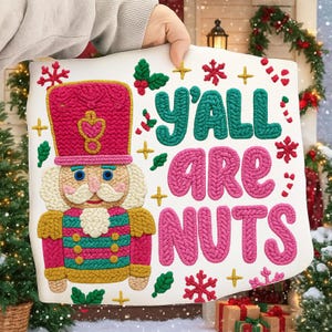 May include: A white decorative sign featuring a colorful nutcracker design and the text "Y'ALL ARE NUTS." The sign has a pink and red hat, a blue-eyed face, and a green, pink, and gold uniform. Festive holiday decor.