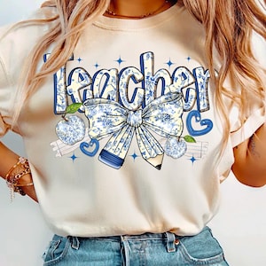 May include: A white t-shirt with the word "teacher" printed in blue and white floral lettering. The design includes a blue and white floral bow, a pencil, and apples.
