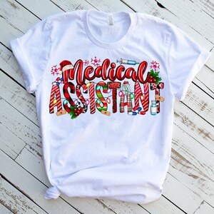 Puede incluir: Camiseta blanca con un dise&ntilde;o navide&ntilde;o que dice "Asistente M&eacute;dico". El dise&ntilde;o incluye bastones de caramelo, luces y s&iacute;mbolos m&eacute;dicos.