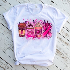 May include: White t-shirt with a pink ribbon design featuring four coffee cups with pink butterflies and pink splatters.