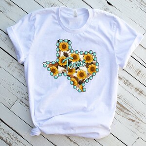 Puede incluir: Camiseta blanca con un gr&aacute;fico del estado de Texas delineado con cuentas turquesa y relleno con un patr&oacute;n de estampado de vaca y girasoles.