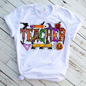 May include: White t-shirt with a Halloween-themed "Teacher" graphic. The graphic features a witch's hat, a spider web, a pumpkin, a pencil, a ruler, and a crow.