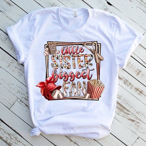 K&ouml;nnte beinhalten: Wei&szlig;es T-Shirt mit einem Grafikdesign eines Baseball-Themas. Der Text auf dem Shirt lautet "Little Sister Biggest Fan".