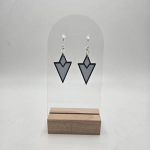 May include: A pair of geometric earrings with a gray interior and black outline, suspended from silver-colored hooks. The earrings are displayed on a clear acrylic stand with a wooden base, set against a white backdrop.