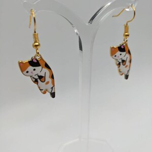 Könnte beinhalten: Ein Paar goldfarbene Ohrringe in Katzenform. Jeder Ohrring hat ein Cartoon-Katzen-Design mit orangefarbenem, schwarzem und weißem Emaille. Die Ohrringe hängen an goldfarbenen Fischhaken-Ohrdrähten. Die Ohrringe werden auf einem transparenten Acrylständer ausgestellt.
