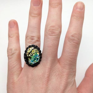 Può includere: Un anello nero con una pietra ovale in vetro iridescente. La pietra mostra sfumature di verde, blu e giallo. L'anello ha un bordo decorativo in filigrana nera e viene indossato su un dito.