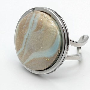 Pode incluir: Um anel prateado com uma pedra grande e redonda marmorizada. A pedra apresenta redemoinhos de bege, azul claro e dourado. O anel tem uma banda larga e um design aberto único.