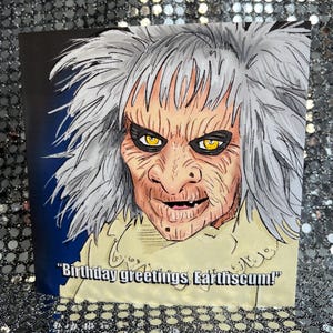 Puede incluir: Tarjeta de felicitación con una ilustración detallada de un personaje con pelo gris, ojos amarillos y una expresión severa. La tarjeta incluye el texto "Birthday greetings, Earthscum!" en blanco.