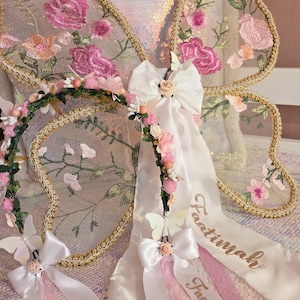 May include: A pair of sheer butterfly wings featuring pink floral embroidery and gold trim, paired with a floral crown headband. The headband includes white satin bows, small pink flowers, and long white ribbons embroidered with the name Faatimah.