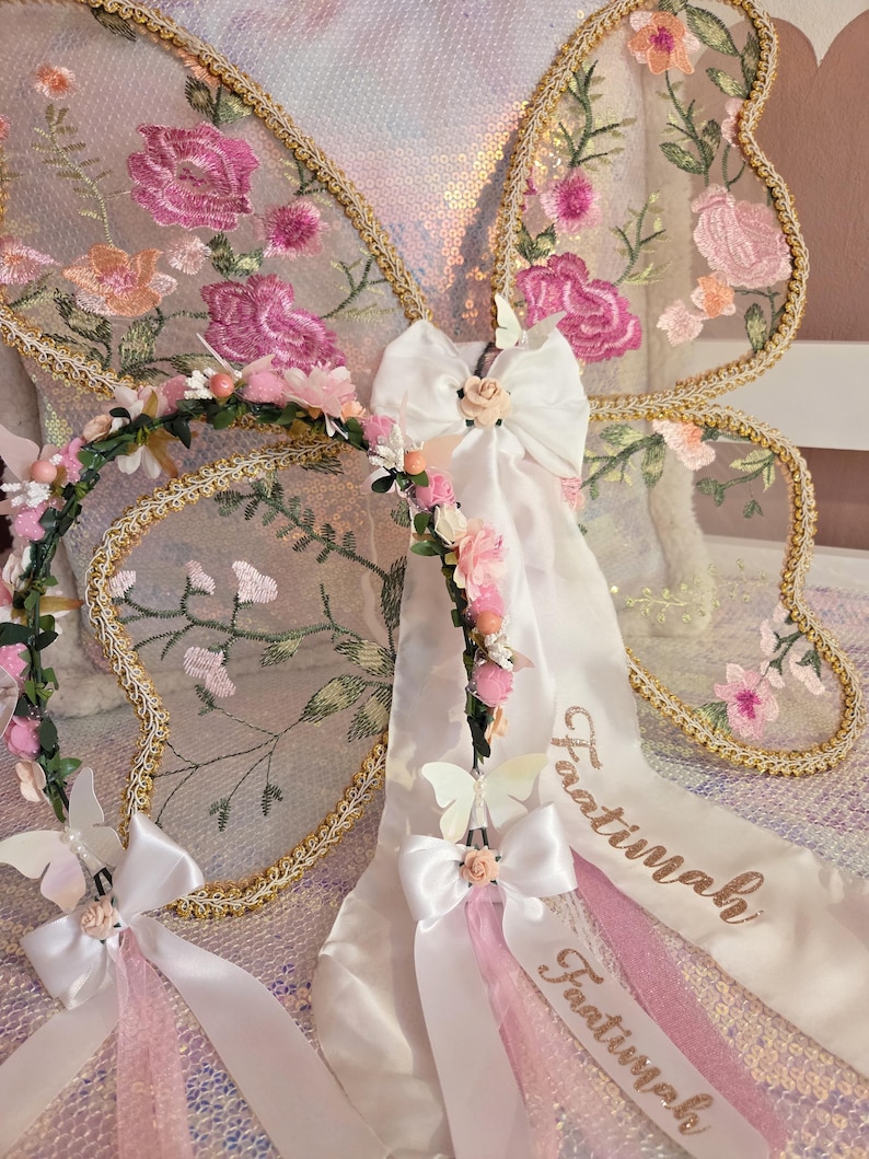 May include: A pair of sheer butterfly wings featuring pink floral embroidery and gold trim, paired with a floral crown headband. The headband includes white satin bows, small pink flowers, and long white ribbons embroidered with the name Faatimah.