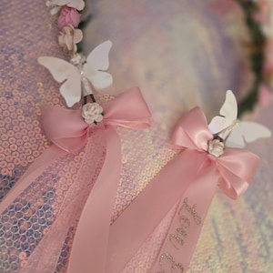 May include: A floral crown featuring pink and white fabric flowers, accented with white butterfly cutouts and pink satin ribbons. One ribbon displays the name Lou Ella in gold glitter script. The accessory is adorned with small white roses.