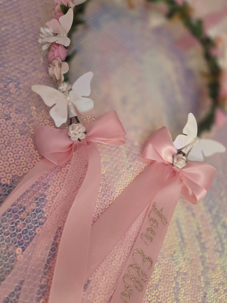 May include: A floral crown featuring pink and white fabric flowers, accented with white butterfly cutouts and pink satin ribbons. One ribbon displays the name Lou Ella in gold glitter script. The accessory is adorned with small white roses.