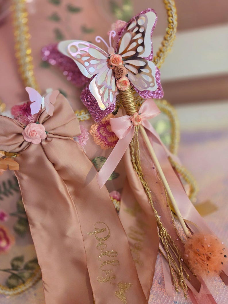 May include: A decorative wand featuring a large iridescent butterfly with pink glitter accents, small peach-colored fabric roses, and long satin ribbons in a dusty rose shade. The wand handle is wrapped in gold cord and includes a small peach bow.