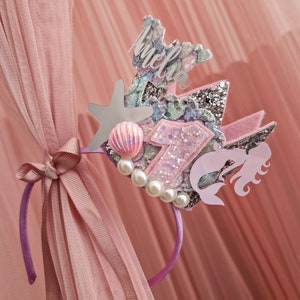 May include: A purple headband with a pink, blue, and silver mermaid crown. The crown features a star, a seashell, and the number "1". The crown also has the name "Amelia" written on it.