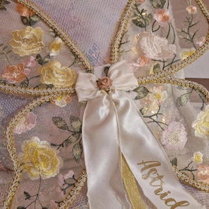 May include: Butterfly wings made of sheer fabric with embroidered yellow and pink floral patterns, edged in gold trim. A cream-colored satin bow with a small rose sits at the center, featuring long ribbons with the name Astrid printed in gold.