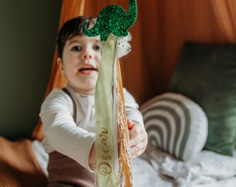 May include: A child holds a green glitter dinosaur wand with a yellow, white, and brown ribbon. The ribbon has the word "Declan" written on it in gold glitter.