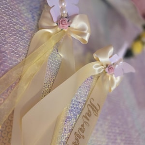 May include: Two decorative wands featuring iridescent sequin fabric, cream-colored satin ribbons, and a light purple butterfly accent. Each wand is topped with a small pink rose and white pearls. The name Nalani is written in gold glitter on one ribbon.