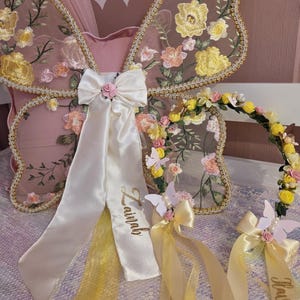 May include: A set of butterfly wings and a floral crown. The wings feature sheer mesh with embroidered yellow and peach flowers and gold trim. A white satin bow with the name Zainab is attached. The crown has yellow and pink flowers and ribbons.