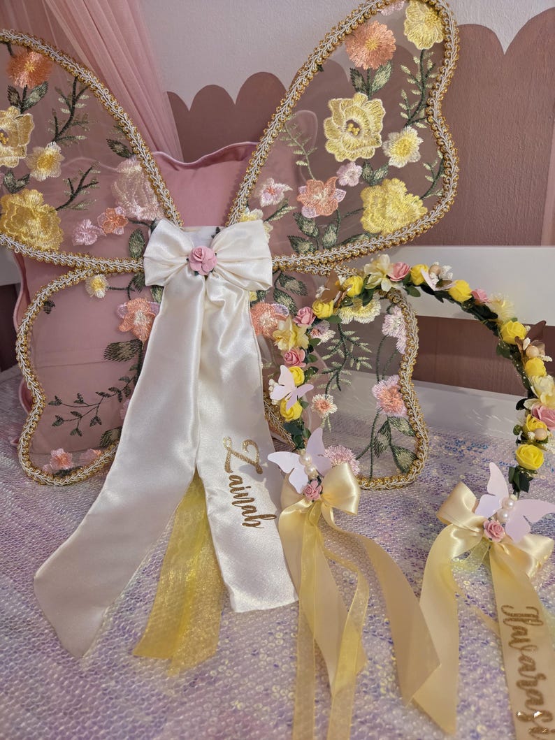May include: A set of butterfly wings and a floral crown. The wings feature sheer mesh with embroidered yellow and peach flowers and gold trim. A white satin bow with the name Zainab is attached. The crown has yellow and pink flowers and ribbons.