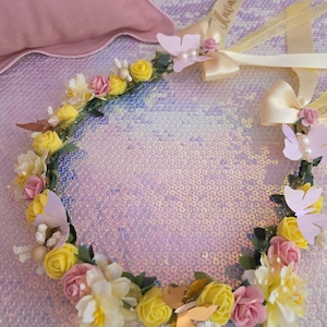 May include: A floral crown featuring yellow and pink foam roses, white blossoms, green leaves, and assorted butterfly accents. The headpiece includes two cream-colored satin ribbons, one of which displays the cursive text "Avarasi".