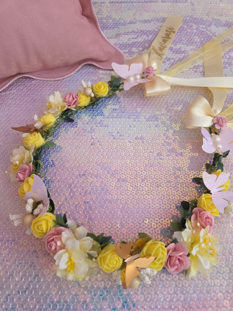 May include: A floral crown featuring yellow and pink foam roses, white blossoms, green leaves, and assorted butterfly accents. The headpiece includes two cream-colored satin ribbons, one of which displays the cursive text "Avarasi".
