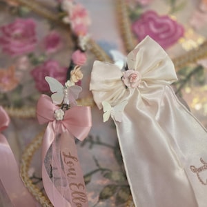 May include: Two decorative hair accessories featuring satin ribbons, small fabric roses, and white butterfly accents. One ribbon is light pink with the name Lou Ella printed in glittery script, while the other is cream-colored with a matching rose center.
