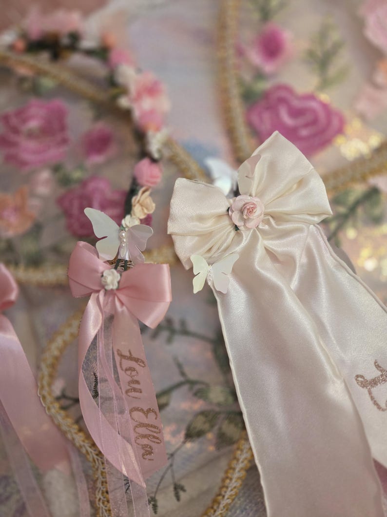 May include: Two decorative hair accessories featuring satin ribbons, small fabric roses, and white butterfly accents. One ribbon is light pink with the name Lou Ella printed in glittery script, while the other is cream-colored with a matching rose center.