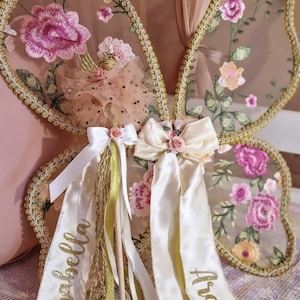 May include: A pair of butterfly wings featuring sheer fabric with embroidered pink and orange floral patterns and gold trim. A small doll with a tulle skirt is attached, adorned with white satin ribbons featuring the name Arabella in gold script.