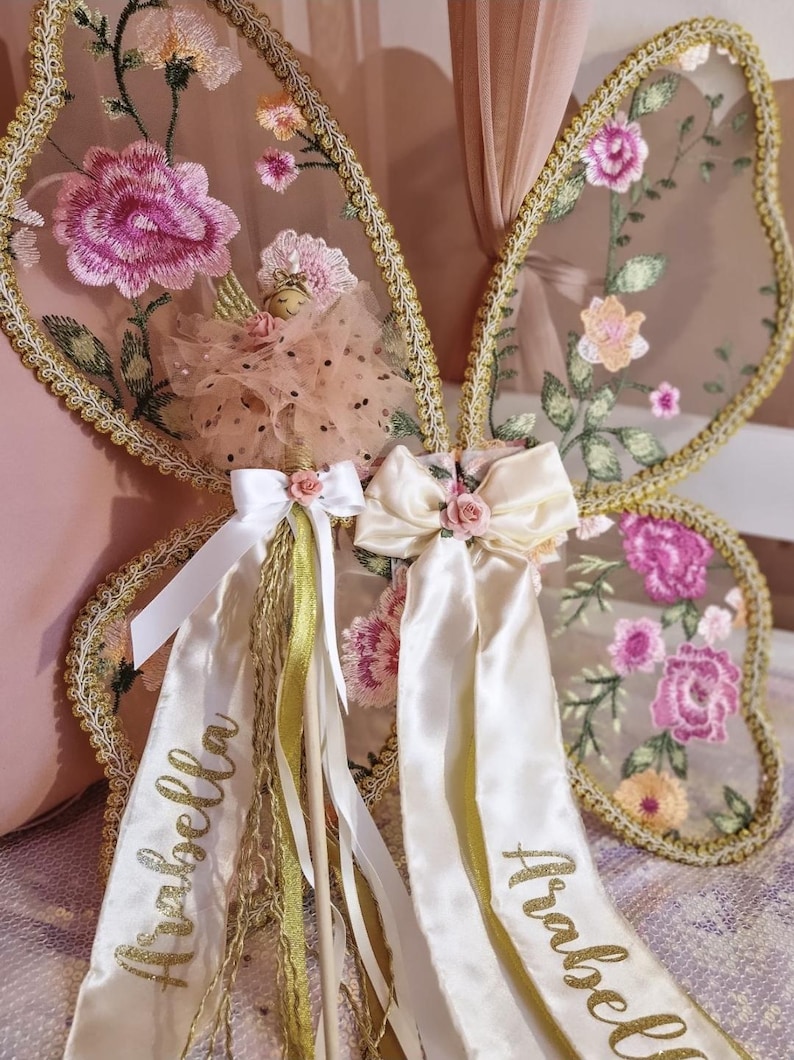 May include: A pair of butterfly wings featuring sheer fabric with embroidered pink and orange floral patterns and gold trim. A small doll with a tulle skirt is attached, adorned with white satin ribbons featuring the name Arabella in gold script.
