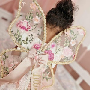 May include: Butterfly wings featuring sheer fabric with embroidered pink and white floral patterns, gold trim edges, and a satin ribbon bow at the center. A long ribbon tail hangs down with the name Lillia embroidered in gold glitter script.