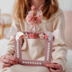 May include: A pink picture frame with a ballerina figurine on top, decorated with pink flowers and white butterflies. The frame is surrounded by a row of white pearls.