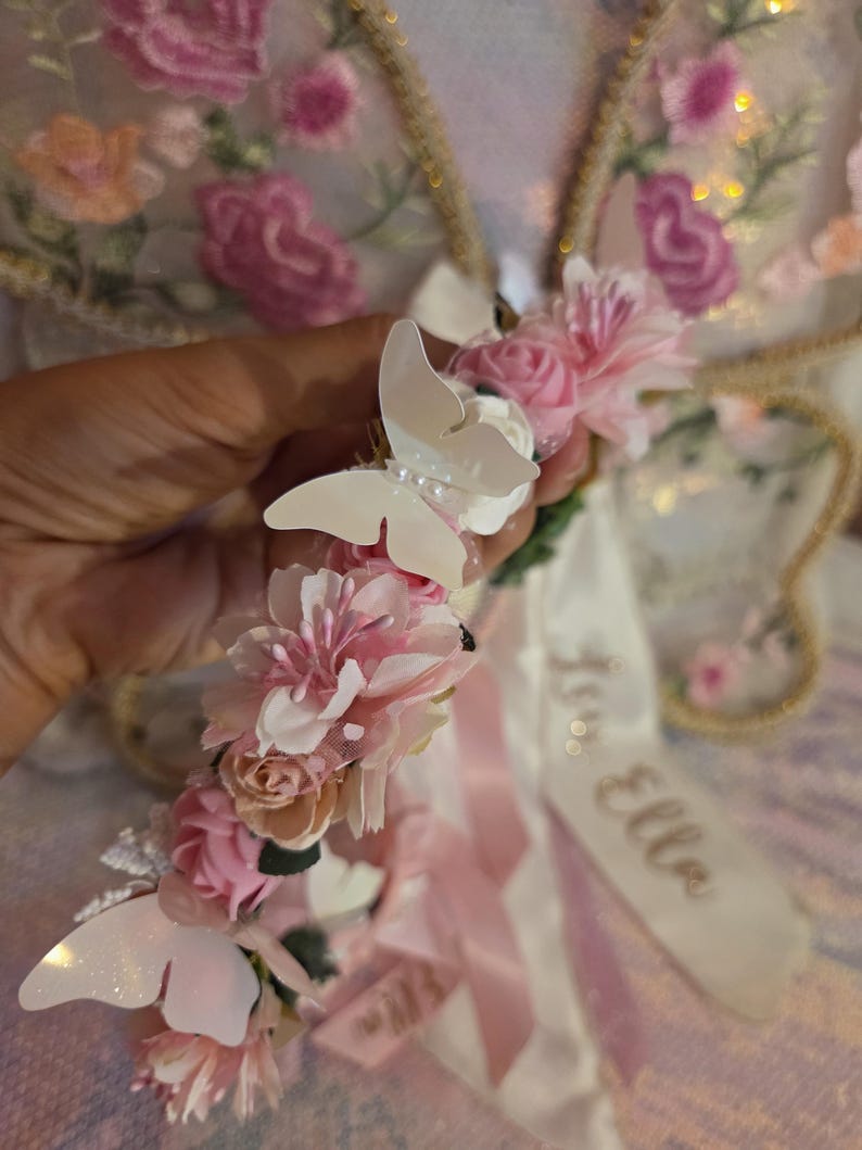 May include: A floral headband featuring pink fabric roses, white artificial flowers, and white butterfly accents. A white satin ribbon hangs from the headband with the name Ella printed in gold script. The design includes small pearl-like beads.