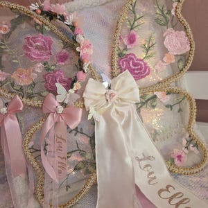 May include: Butterfly wings featuring sheer fabric with pink and peach floral embroidery, gold trim, and a cream satin bow. A matching floral headband and satin ribbons are attached, with the name Lou Ella embroidered in gold script on the ribbons.