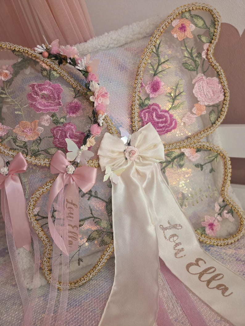 May include: Butterfly wings featuring sheer fabric with pink and peach floral embroidery, gold trim, and a cream satin bow. A matching floral headband and satin ribbons are attached, with the name Lou Ella embroidered in gold script on the ribbons.