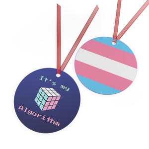 May include: Two round ornaments with red ribbon. One ornament is blue with a pixelated Rubik's cube and the text "It's my Algorithm". The other ornament is a blue, pink, and white transgender pride flag.