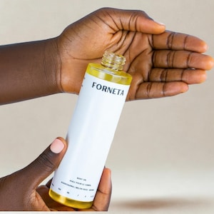 May include: A cylindrical body oil bottle with a white label and the word "FORNETA" in black. The bottle is held in a person's hands. The bottle's contents are a yellow liquid. The label also includes the words "BODY OIL" and "HUILE POUR LE CORPS".