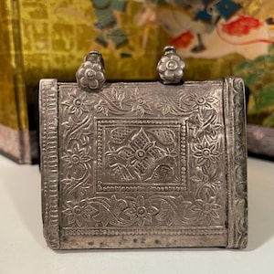 Könnte beinhalten: Ein silberfarbener, rechteckiger Anhänger mit aufwendigen floralen und geometrischen Mustern. Zwei kleine, blütenförmige Schlaufen befinden sich oben. Die Oberfläche des Anhängers ist mit geprägten Designs versehen, die einen Vintage-Look zeigen.