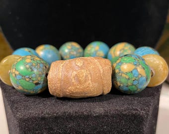 Puede incluir: Un brazalete hecho de cuentas turquesa y marrones. El brazalete tiene una cuenta grande de color marrón en el centro con una imagen tallada de una persona sentada en una postura meditativa.
