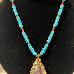 May include: A turquoise and gold beaded necklace with a gold pendant featuring a small, framed image of a person. The pendant is decorated with colorful enamel.