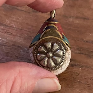 May include: A white shell pendant with a silver flower design and a colorful, multi-layered top. The top is made of red, green, blue, and gold metal.